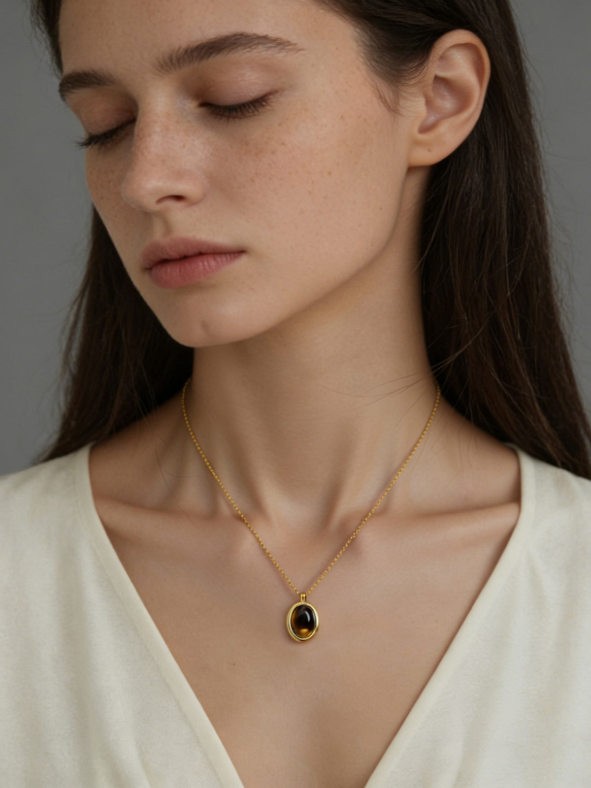 Oval Glow Pendant Necklace in gold with a brown stone, shown on a model with eyes closed, front view